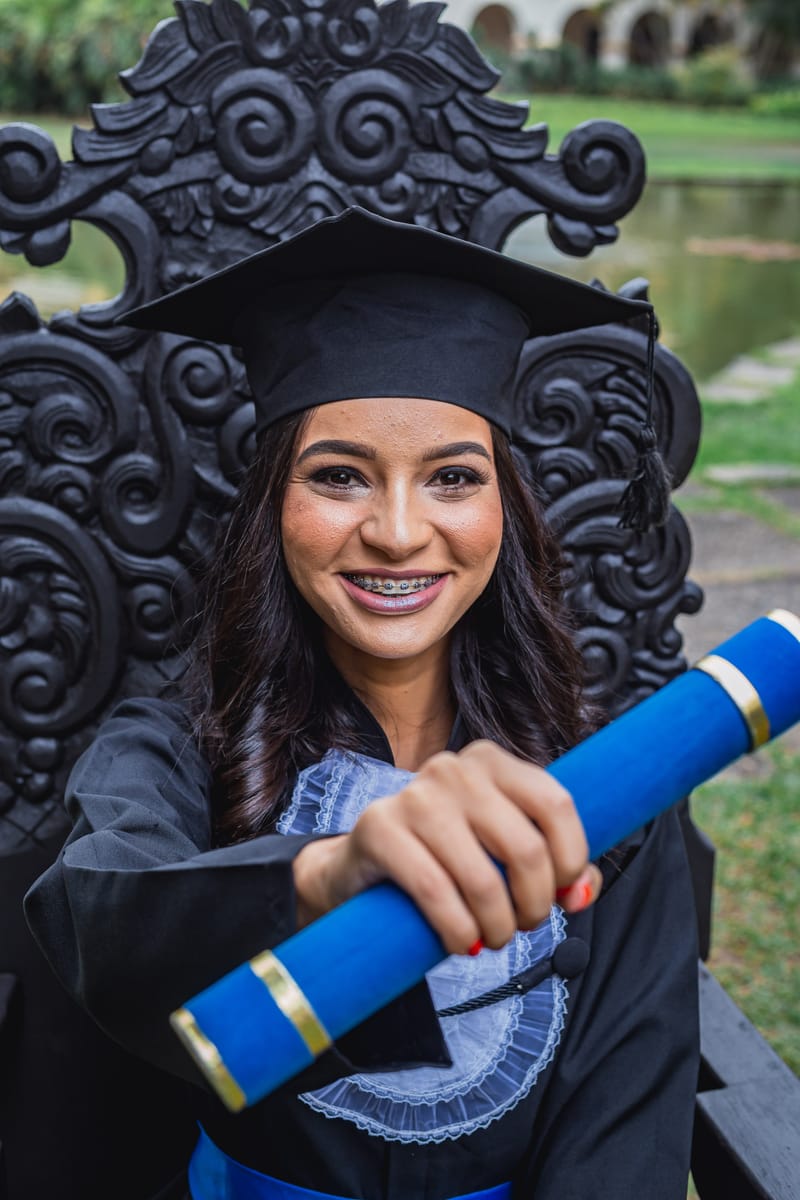Formanda sorrindo na cadeira de formatura, estendendo o diploma