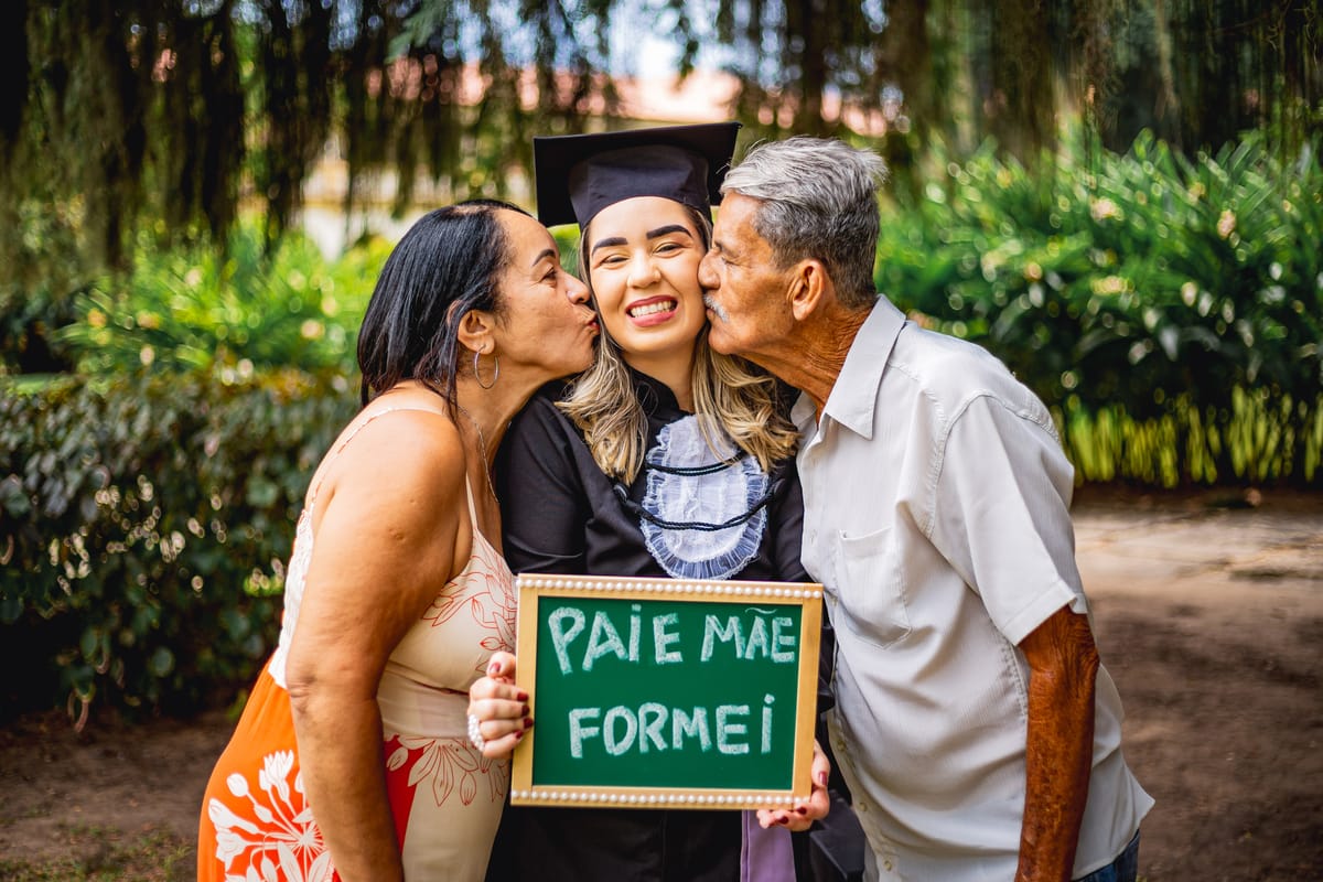 Formanda recebendo beijo dos pais, com a placa “Pai e mãe, formei!”